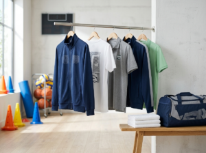 Hochwertige Vereinsausstattung mit bedruckten Trainingsjacken, T-Shirts und Polos auf einer Kleiderstange sowie einer Sporttasche in einer modernen Sportumgebung